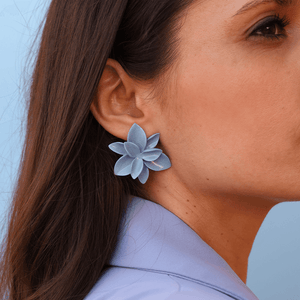 Zoom-in: porcelain floral earrings. Feminine and delicate jewelry. Petal-inspired blue earrings are being worn by a brunette model. She is also wearing a blue blazer, matching the blue background.