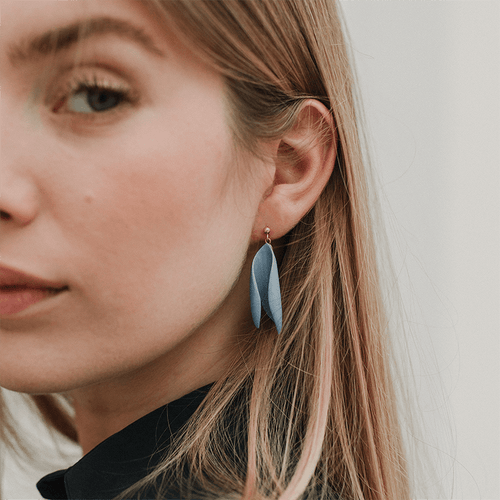 Woman looking straight into the camera. She's blond and is wearing timeless earrings made of light blue porcelain.